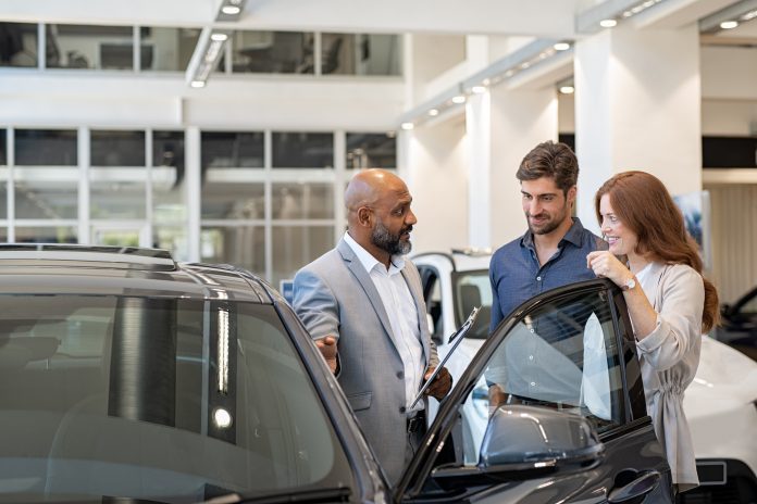 Mature salesman delivering an excellent customer experience to a man and woman in car dealership showroom.