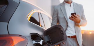 6 essential pillars of mainstream electric vehicle adoption A man holding smartphone while charging car at electric vehicle charging station, closeup.A man holding smartphone while charging car at electric vehicle charging station, closeup.
