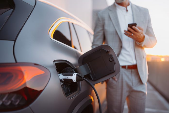 A man holding smartphone while charging car at electric vehicle charging station, closeup.A man holding smartphone while charging car at electric vehicle charging station, closeup.